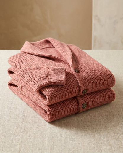 Two terracotta-pink ribbed cardigans folded in a stack on a linen-covered table, showing the shawl collar, buttons and ribbed cuff. 7