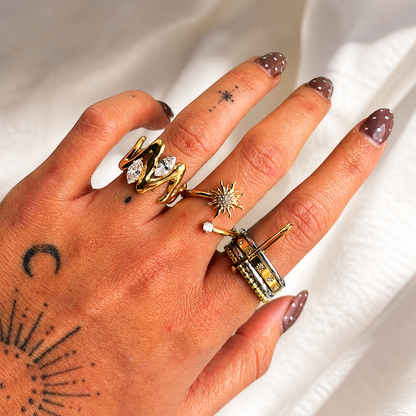 Hand wearing the wavy gold-tone ring with clear stones, stacked alongside other rings, against white fabric. 3