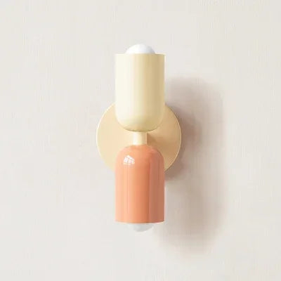 Single wall lamp with a cream upper shade and backplate and a peach lower shade, shown against a plain pale wall. 16