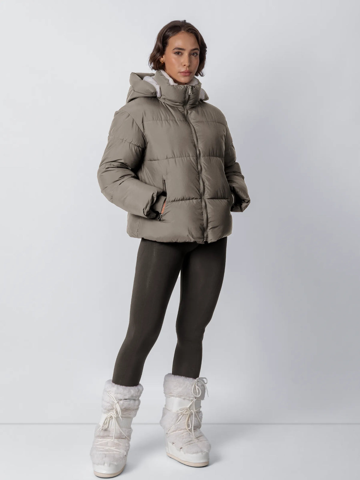 Model wearing the taupe puffer jacket zipped up with hood on, dark leggings and white fluffy snow boots 21