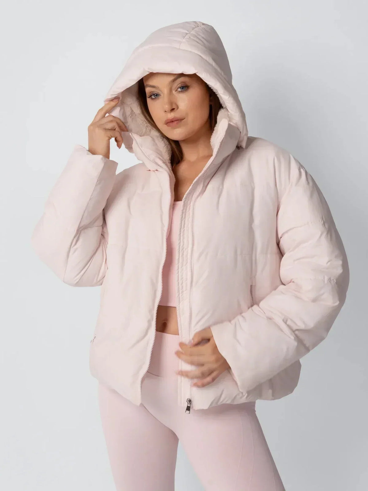 Model in pale pink puffer jacket with the padded hood up, one hand holding the hood edge 6