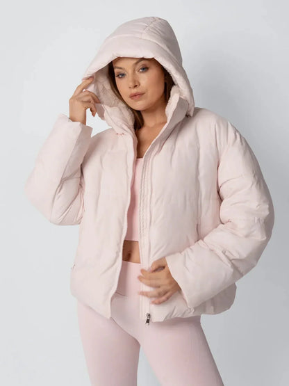 Model in pale pink puffer jacket with the padded hood up, one hand holding the hood edge 6