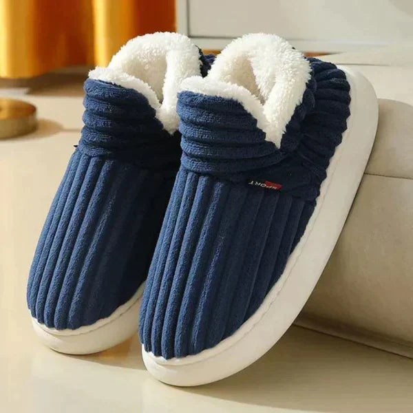 Pair of navy blue ribbed plush slippers with white fleece lining and thick white soles, resting against a sofa. 1
