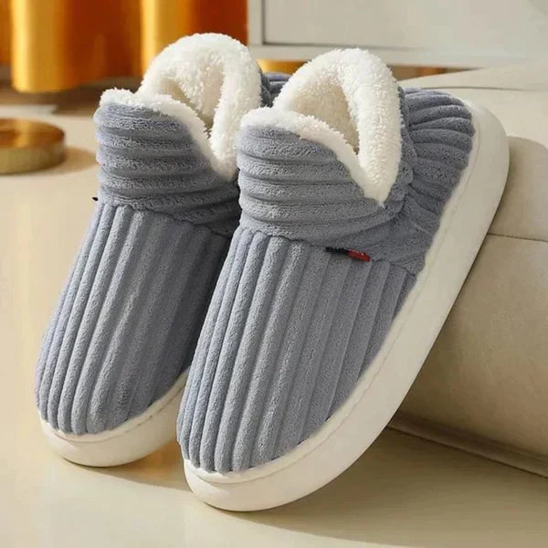 Pair of grey ribbed plush slippers with soft white fleece collars and thick cream soles, shown beside a sofa. 2