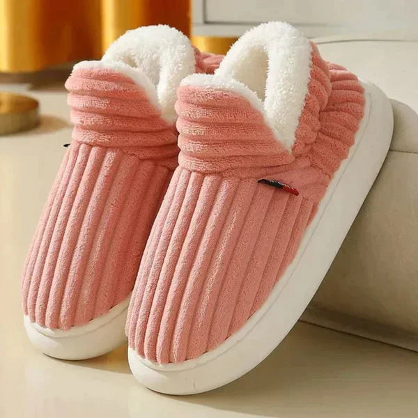 Pair of coral pink ribbed plush slippers with white fleece lining and thick white soles, set beside a sofa. 5