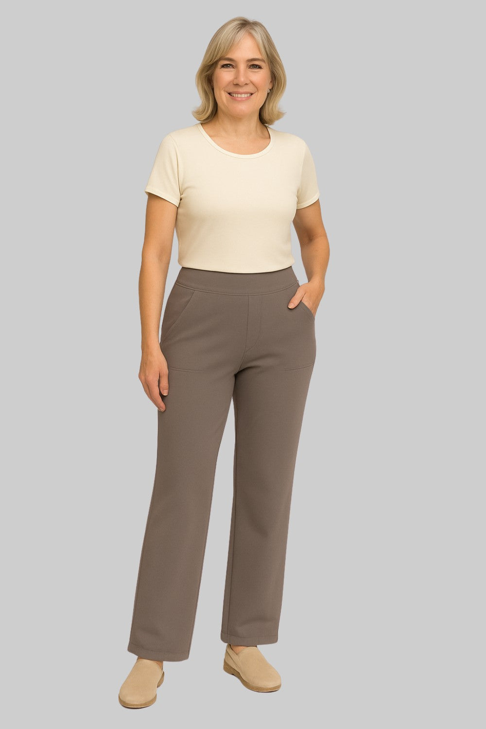 Smiling woman wearing taupe trousers with a cream short-sleeved top and beige slip-on shoes, hand resting in pocket. 2