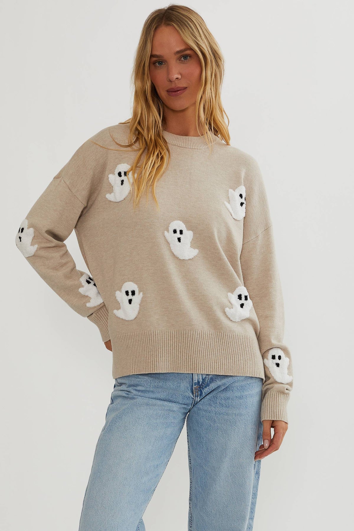 Woman with long blonde hair wears the oatmeal knit jumper with fluffy white ghost appliqués, ribbed hem and cuffs, and light blue jeans. 1