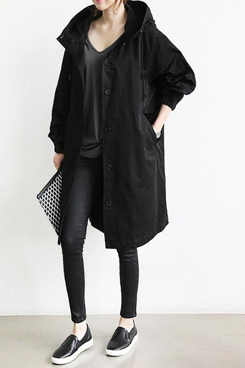 Black hooded button-front coat worn open over a dark top, leggings and slip-on shoes, with a patterned clutch 2