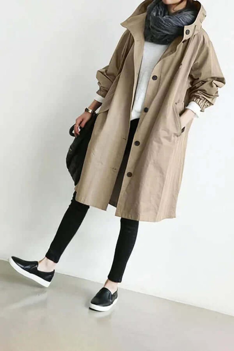 Camel-coloured coat worn with black leggings and slip-on shoes, leaning against a wall, carrying a black bag 5