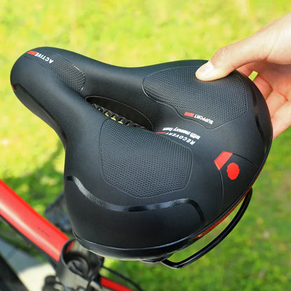 Hand holding the black saddle outdoors, showing the textured pads, central cutout, red accents and metal rails beneath. 5