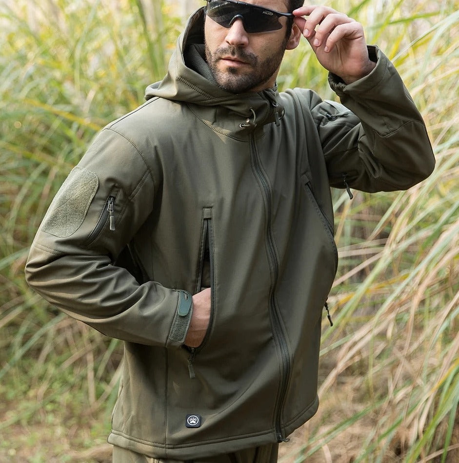 Man in olive green hooded jacket and sunglasses standing in tall grass, one hand in the zipped front pocket. 11