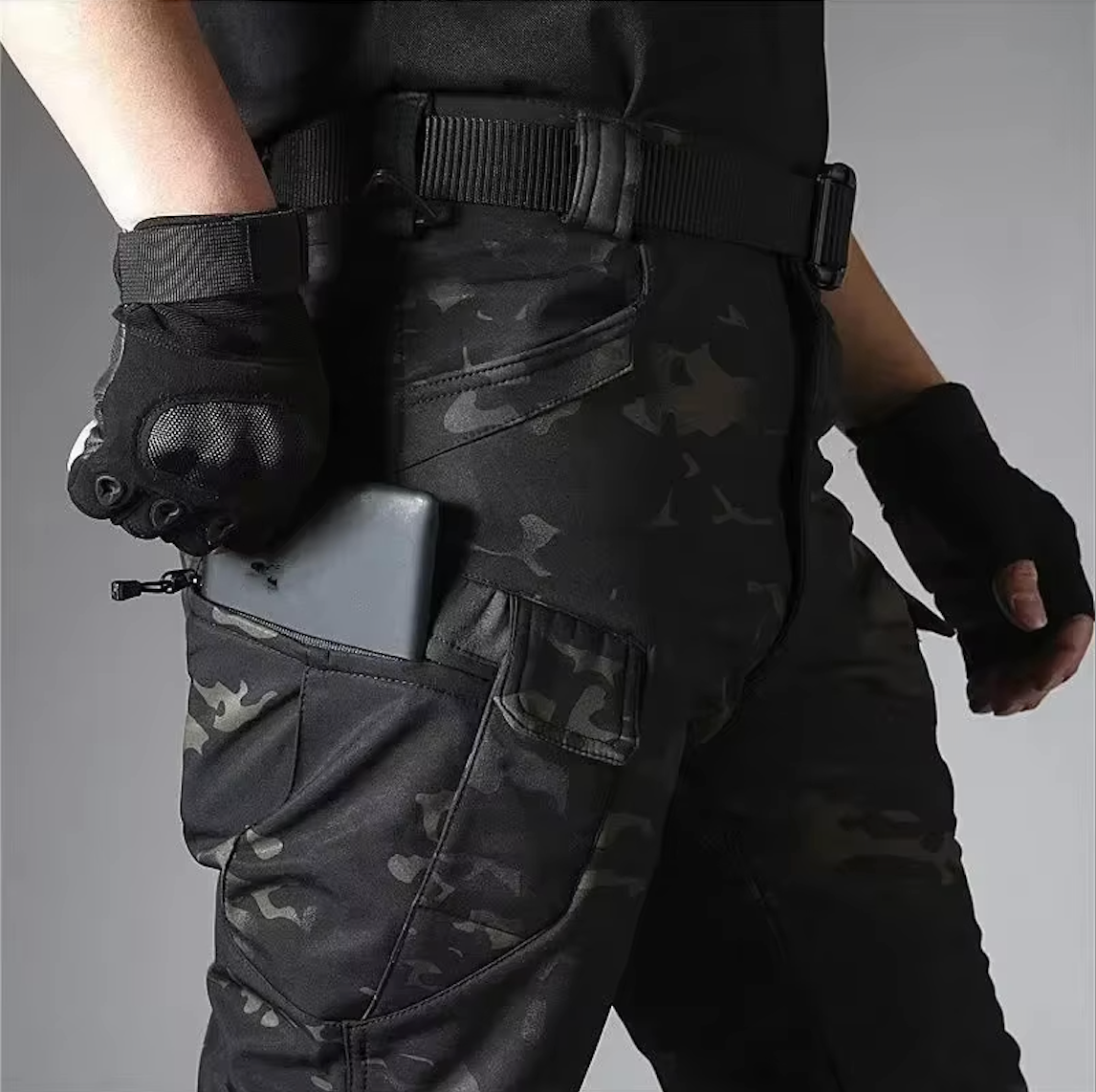 Detail of dark camouflage trousers worn with a belt, a phone being slipped into the zipped thigh pocket. 19